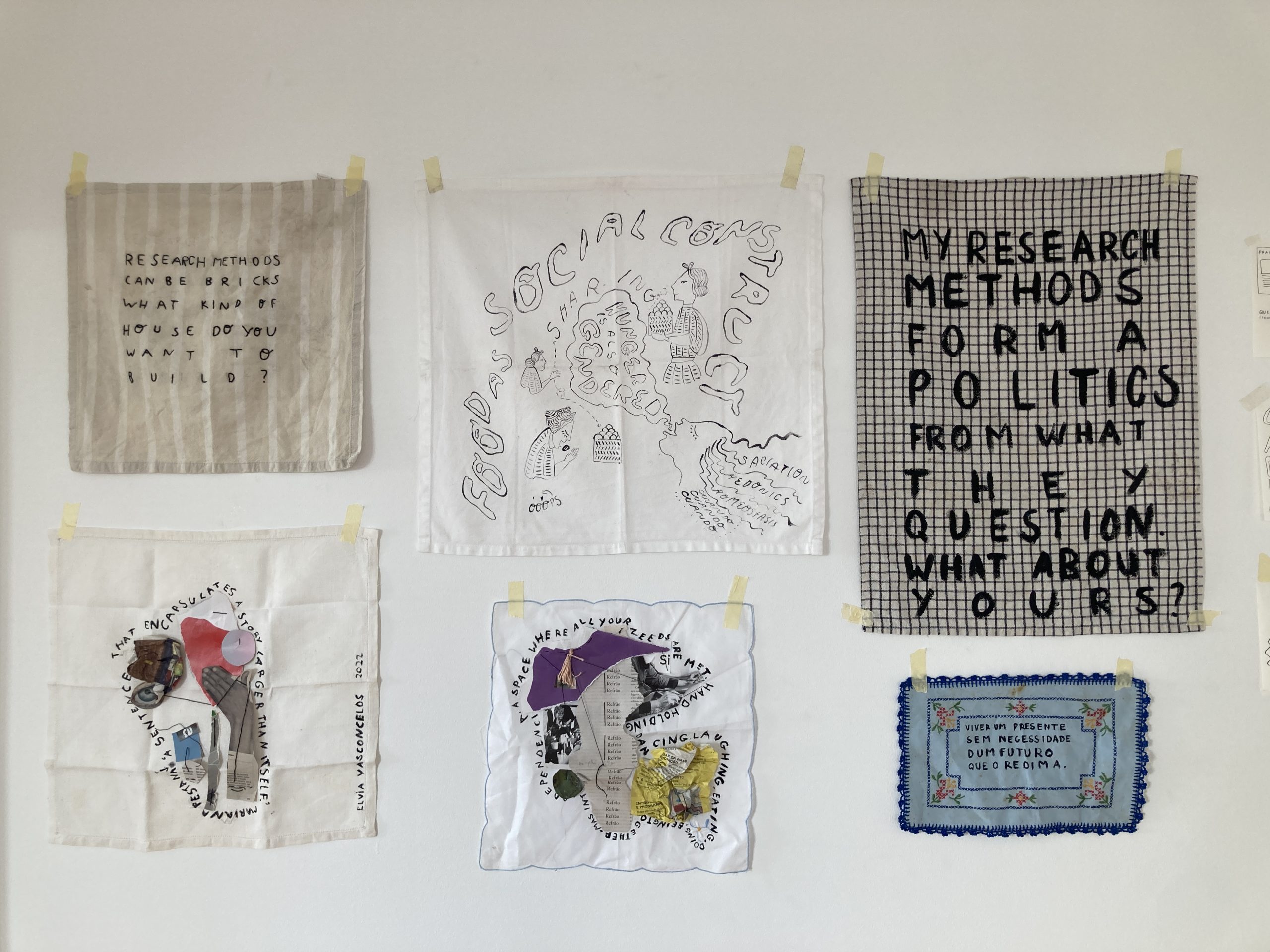 Six pieces of fabric are shown hanging on a white wall. From top left, the first piece is a grey fabric 
               with white stripes, with the following sentence embroidered 'Research methods can be brics. What kind of 
               house do you want to build?'. The second piece, on the top middle row is a white cotton napkin that has 
               been painted with black ink. It reads in wavy bulky outlined letters'Food as a social construct' and shows 
               four figures holding and extending food. One of these figures is annotated with a speech bubble coming from 
               their open mouth that reads 'Hunger is also gendered'. This same figure's wavy hair is annotated with the words 
               saciation, hedonics, homeostasis, and cuando cuando cuando. The third pice on the top row, on the right far corner is 
               a black and white squared patterned kitchen cloth with the sentence 'my research methods form a politics from whaty 
               they questions. What about yours?' painted with black in. On the bottom row, the first piece on the left shows a 
               mixed media collage on a white kitche cloth and is annotated by the following sentence surrounding the collage:
               'A sentence that encapsulates a story larger than itself. Mariana Pestana.' Next to it,in the middle bottom row, 
               there's another mixed media collage with the sentence: 'A space where all your needs are met. Hand holding, dancing,
               laughing, eating, doing, being, together. Mas interdependencia.' The final piece, bottom-left, is a rectangular light
               blue little table cloth, with blue embroidery trimmings, and flowers. The sentence 'Viver um presente sem necessidade
               dum futuro que o redima' (translation from portuguese: To live a present without the need of a future to redeeem it)
               in black ink.